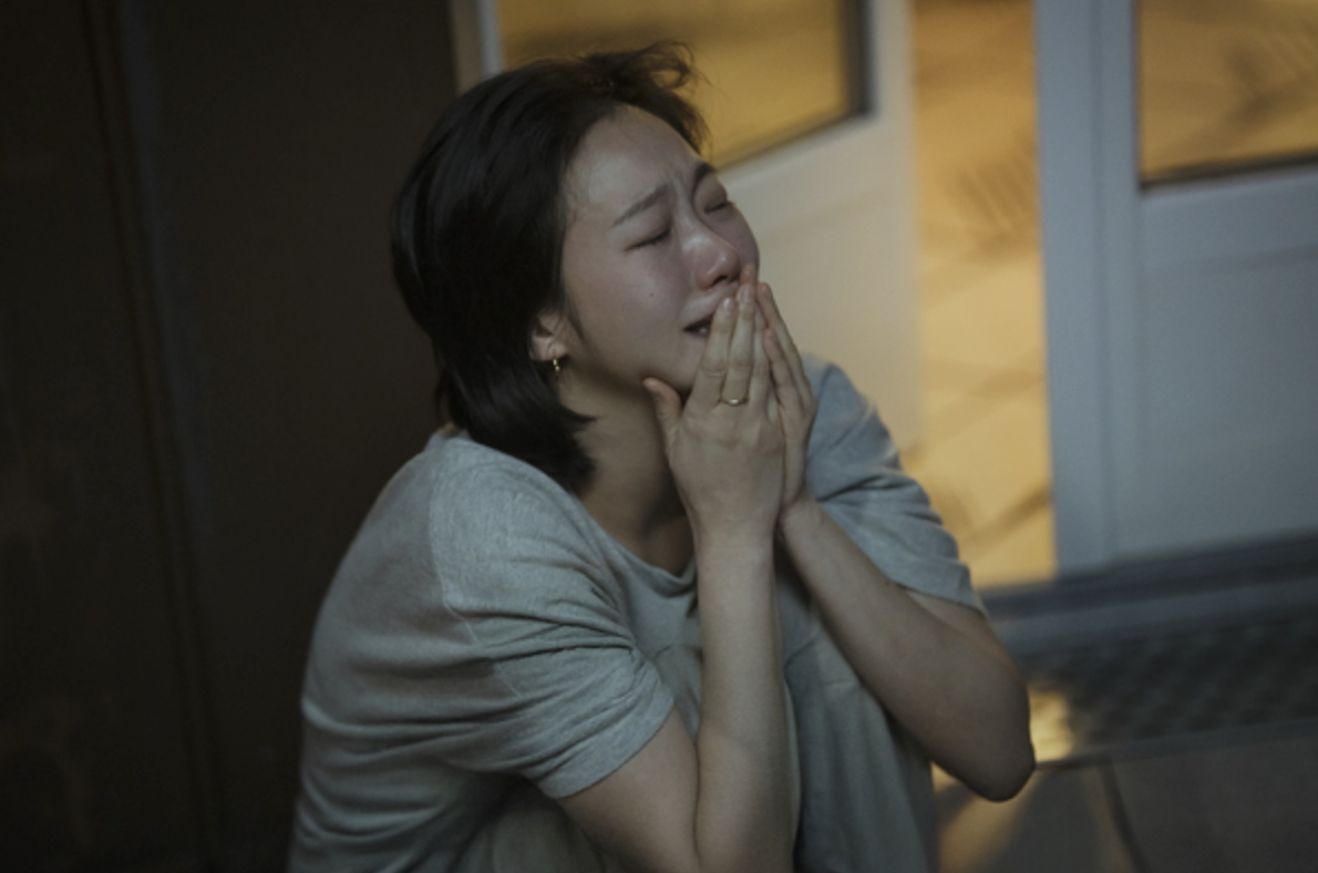 어두운 실내, 유리문 앞에 웅크려 앉은 여성이 두 손으로 입과 코를 감싼 채 눈을 감고 눈물을 흘리고 있다