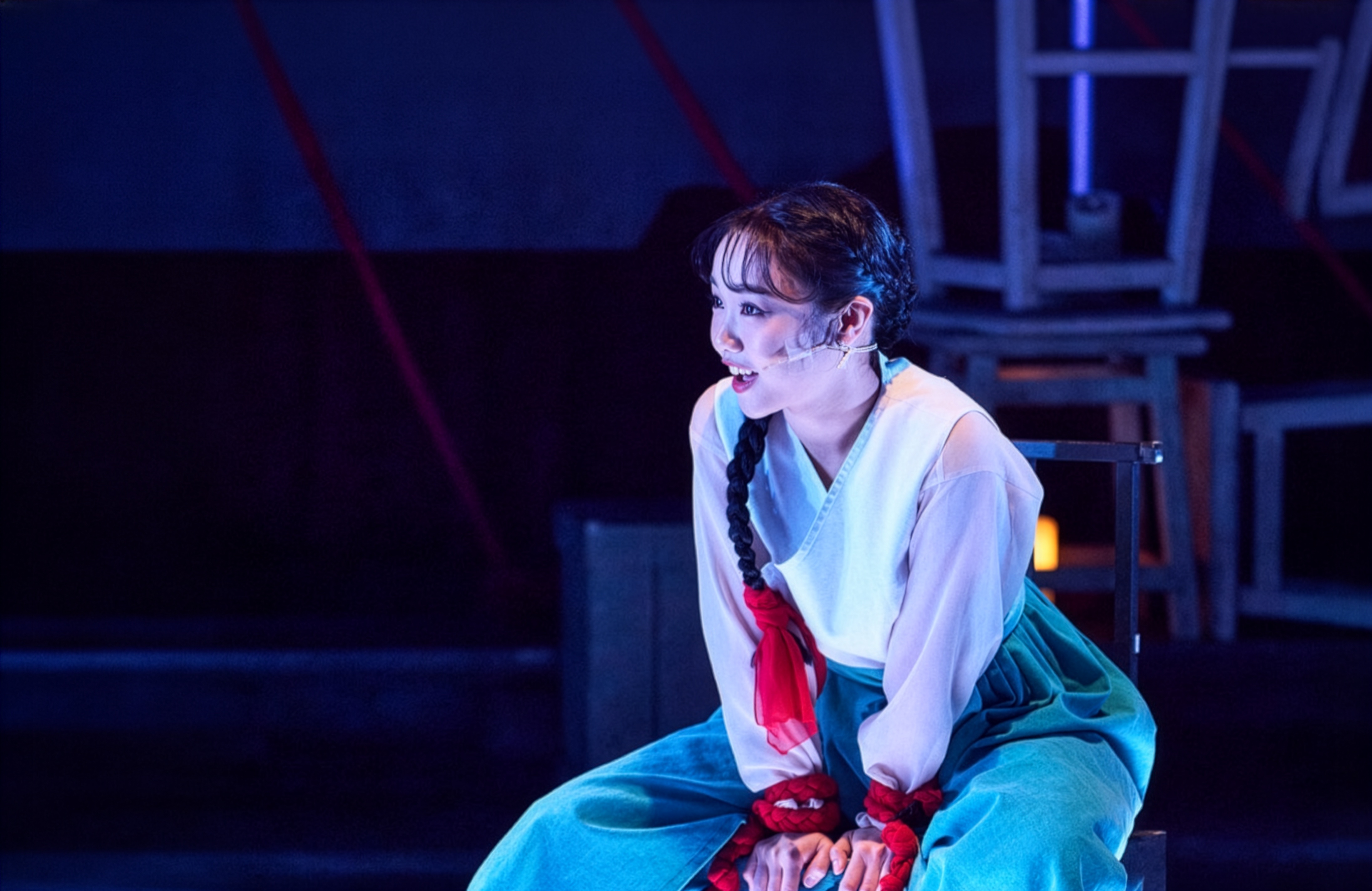 뮤지컬 《홍련》의 한 장면 — 푸른 조명 아래 흰 저고리에 붉은 고름, 옥빛 치마를 입은 홍련이 긴 댕기머리를 하고 두 손이 붉은 포승줄에 묶인 채 입을 벌려 말하고 있다
